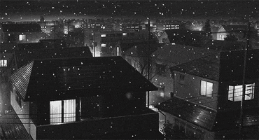 Anime gif. Snow falls gracefully over a quiet town with houses lit from inside. There is a nostalgic ambience to the black and white scenery.