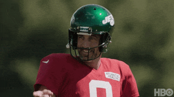Sports gif. Coach Aaron Rodgers in a red jersey and green Jets helmet gives someone offscreen a cheesy smile as he raises a finger to point at them. 