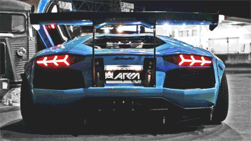 Video gif. From behind, we see a blue Lamborghini as sparks fly from its mufflers.