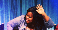 Video gif. A smiling woman raises a hand above her head, pointing one finger up. Text, “this.”