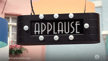 TV gif. From The Good Place, a black art-deco-style electronic sign rocks gently back and forth and reads "Applause" in flickering white text bordered by small white bulbs.