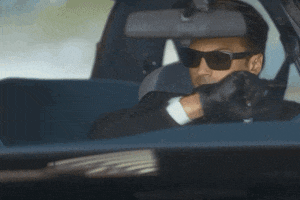 Movie gif. Jonathan T. Baker as the Agent wears sunglasses and sits in the driver’s seat of a car. He looks over with a serious expression, out of the passenger seat window and points at someone in another car beside him.