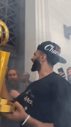 Sports gif. Jamal Murray of the Denver Nuggets wears his team's championship hat shaking a giant basketball trophy and cheering at a group of fans and bystanders. One fan responds by flexing his biceps and yelling back excitedly at Murray. 