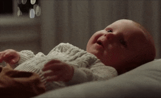 Video gif. A baby in a sweater smiles happily.