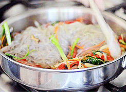Video gif. Closeup of a steaming pan of japchae as a spatula folds in the noodles.
