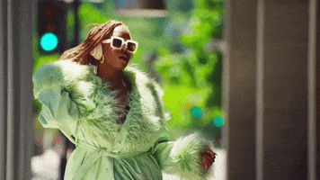 Music video gif. From the video for "DND," Zyah Belle flips her hair as she walks unbothered down a sidewalk, dressed in a mint-colored fuzzy-edged robe and wearing white headphones and sunglasses.