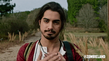 Movie gif. Avan Jogia as Berkeley from Zombieland: Double Tap stands on a grassy path, smiling at us as he tenderly puts his hands over his heart.