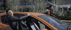Music video gif. From a video by 99 Percent, a guy leans out of an orange car with a wide-eyed and wide-mouthed expression of shock, while another guy standing by the driver's side adjusts his jacket.