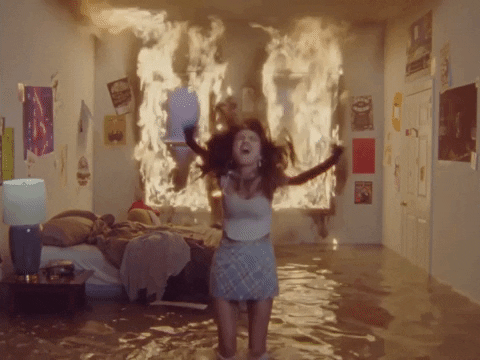A video gif from the Olivia Rodrigo music video Good 4 U. It features a young woman jumping and scream-singing wearing a short preppy skirt and pink babydoll t-shirt with long black gloves. She's standing in a flooded living room where the curtains are on fire. It's the epitome of pop teen anger.