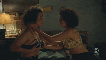 TV gif. Alia Shawkat, as Adele on Broad City, sits on the edge of the bed and pulls in Ilana Glazer as Ilana for a kiss, and the two fall sideways onto the bed.