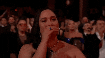 Oscars 2024 GIF. Lily Gladstone, standing in ovation, cups her hand to discreetly cheer a performer intimately.