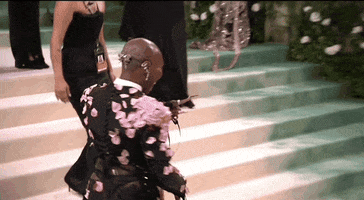 Met Gala 2024 gif. Cynthia Erivo and Ariana Grande move across the carpet for a hug and cheek kiss. The back of Erivo's Thom Browne deconstructed tuxedo features a low rise skirt that shows the top of a black thong. Grande is wearing a pale pink Loewe gown with a corset-style bodice and flowing pleated skirt.
