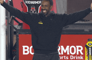 Video gif. Wilfried Nancy, head coach of Columbus Crew, stands on the sidelines, sweeping his arms out and bringing them back in to clap enthusiastically as he grins widely. 