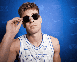 Video gif. Dallin Hall, a basketball player for Brigham Young University, pulls his sunglasses down to the edge of his nose to look at us with a sassy, questioning expression against a blue background covered in sports insignias.