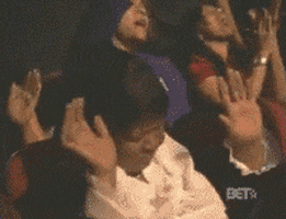 Video gif. Seated in an auditorium full of people applauding, a woman seems to be having a spiritual moment as she holds both hands up and sways with her eyes closed. Without opening her eyes, she starts clapping along.