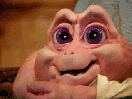 TV gif. Baby Sinclair on the 1990s show Dinosaurs sits in someone’s lap and looks at us with a shocked look, biting his lip. He then busts out in a joyful laughter.