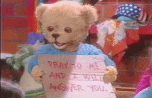 Video gif. A teddy is smiling and nodding its head slightly while holding a sign that says, "Pray to me and I will answer you."