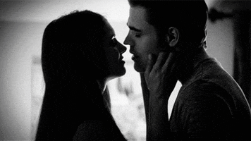 TV gif. A dark black-and-white scene from The Vampire Diaries of a man and woman kissing.