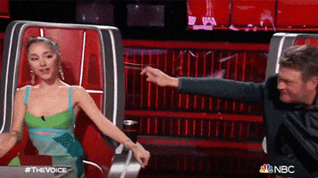 Reality TV gif. Luke Bryan on the Voice reaches out from his chair and points at Ariana Grande, wiggling his finger like a worm. She rolls her eyes and points back at him.