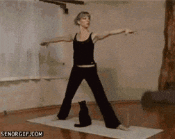 Video gif. Woman stands on a yoga mat with her arms outstretched and her legs spread in a wide triangle; a small cat sits between her legs, then suddenly jumps up and claws the woman's crotch, and the woman yells in pain and grabs herself.
