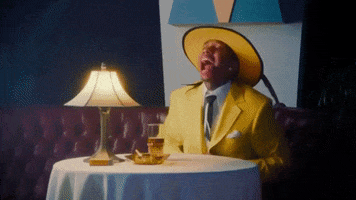 Music video gif. Tyga in Ayy Macarena wears a banana yellow suit and fedora at a table for one. He opens up his suit jacket to allow a heart made out of his white shirt to literally burst out into the open.