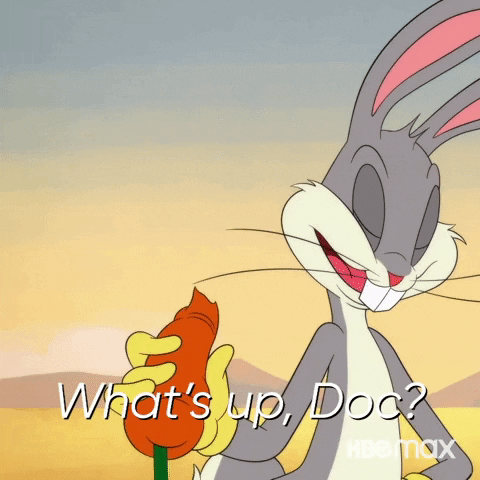 Cartoon gif. A modern Bugs Bunny stands in a desert, holding a carrot with a bite taken out of it as he casually says his classic line. Text, "What's up, Doc?"