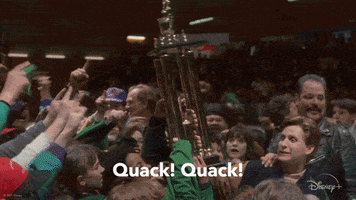 Movie gif. A scene from The Mighty Ducks where they've just won the game and a crowd is on the ice with the team holding the trophy. They cheer simultaneously, "Quack, quack!"