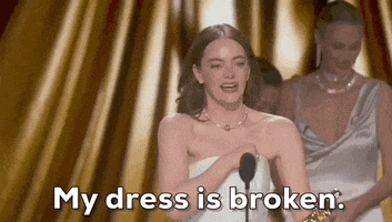 Oscars 2024 GIF. Emma Stone walks up to the microphone while she theatrically turns around to show her bare back and say, "My dress is broken."