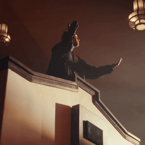 Music video gif. From the video for "Overcompensating," Tyler Joseph of Twenty One Pilots stands at the top of an indoor balcony and has his arms outstretched. He beckons us with both hands, as if saying, "Give it to me."