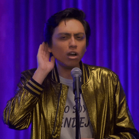 Celebrity gif. Standing on a stage behind a microphone, Martin Urbano puts his hand behind his ear and turns toward the mic gesturing like he can't hear.