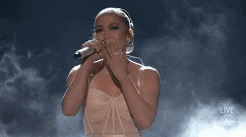 Celebrity gif. On a smoky stage, Jennifer Lopez smiles, blows a kiss to the audience and waves, and bows graciously.