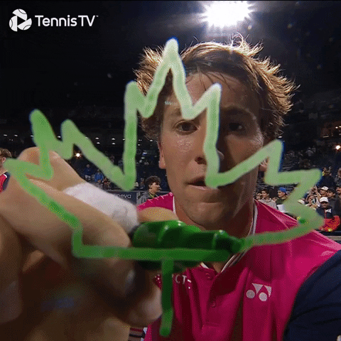 Video gif. Athlete leans in to draw a green maple leaf on the lens of a camera that has been set up to film a tennis match. 