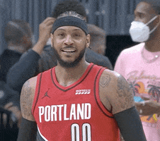 Sports gif. Carmelo Anthony of the Portland Trail Blazers wearing his uniform and looking at us as he claps enthusiastically in front of a few people wearing surgical masks behind him at a game. 