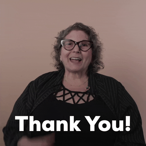 Reaction gif. A Disabled white woman with kinky curly gray hair and big wine-colored cat-eye glasses says "thank you," in ASL and English, punctuating by clutching her heart sweetly.