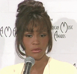 Celebrity gif. Whitney Houston stands at a mic as her eyes go wide in shock before she turns to the side with a confused expression.