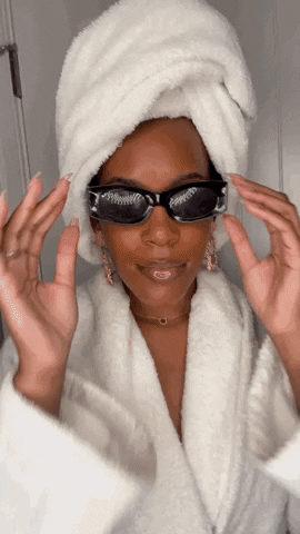 Black woman in a white bathrobe with a white towel wrapped around her hair, she moves forward pulling her black sunglasses down a little to wink.