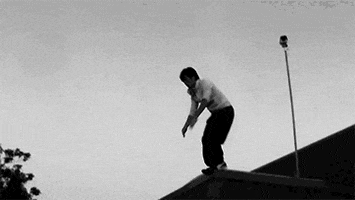Video gif. Black and white footage of a boy practicing parkour, jumping off a roof onto a wall before rolling expertly to the ground.