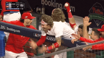 Sports gif. Bryson Stott of the Phillies hangs over the dugout wall and screams in excitement, pumping his arms in a chopping motion as another player hops around on one leg smacking his own rear end. 