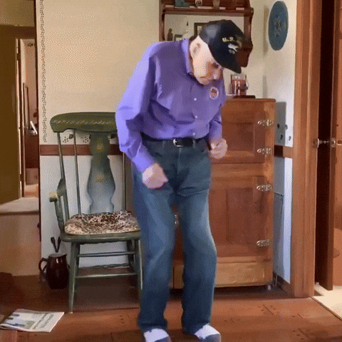 Video gif. Hunched over elderly man wearing white socks and a US Navy hat looks down towards his feet and does a smooth two step dance, sliding his socks along the wood floors as he moves. 