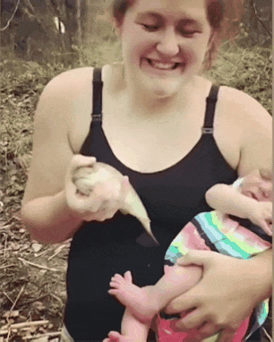 Video gif. A woman is holding her baby in one hand and a fish in the other. The fish jumps out of her hand and flops around on the baby and the camera zooms in on the baby's face who is mad dogging her mom.