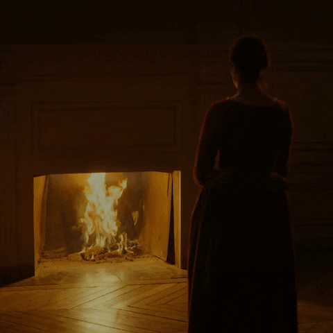 Movie gif. Adèle Haenel as Héloïse in Portrait of a Lady on Fire stands in a dark room as she stares at a fire that roars in a hearth