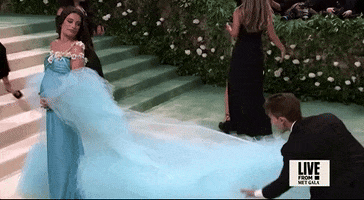 Met Gala 2024 gif. Lea Michele wearing Rodarte, an aqua gown with 3D white flowers across one shoulder and empire waist, and cradling her baby bump, looks on as an assistant fluffs the train her oversized tulle cape worn off her shoulders, then fluffs her hair in slow motion, returning her attention to the cameras.