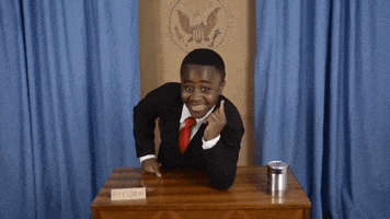 Video gif. Robby Novak as Kid President leans on a desk and points toward us with a big smile.