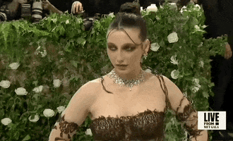 Met Gala 2024 gif. Camera pans up on Emma Chamberlain wearing a gothic-style Jean Paul Gaultier gown with long panels of sheer black fabric that sweep out from her mid back to fall behind her as a train. Dark brown strands of lace are weaved around her arms resembling brambles and ferns.