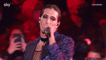 Reality TV gif. Wearing a fishnet shirt with a collar and a sparkly blue jacket, Damiano David from X Factor sings on stage and points out toward us.