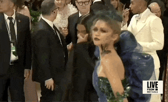 Met Gala 2024 gif. Closeup of the top of Zendaya's midnight blue and dark teal Maison Margiela tulle gown, leaning forward for a couture pose then strutting down the carpet. She wears dark eyeliner and bright red eyeshadow with dark lipstick.