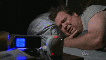 TV gif. Eric Stonestreet as Cam on Modern Family lies in bed in the dark looking at the clock on his bedside table, shaking as he cries. 