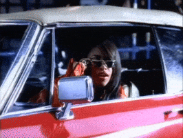 Music video gif. From the video for Got to Give it Up, Aaliyah lowers her sunglasses and peers out the drivers' side window of a red car and smiles.