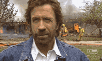 TV gif. Chuck Norris as Ranger Cordell Walker from Walker, Texas Ranger laughs lightheartedly while casually standing in front of a house completely swallowed up in flames, as firefighters work in the background.
