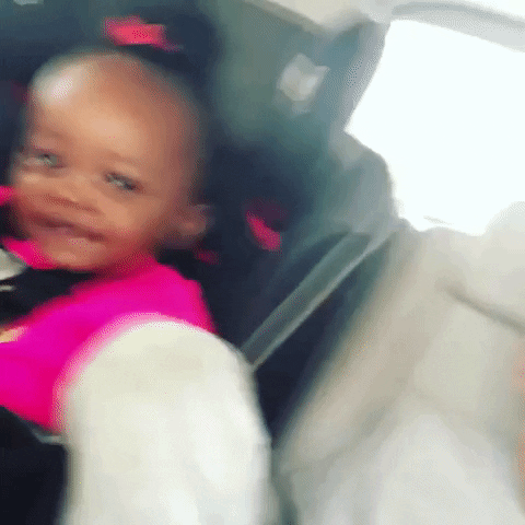 Video gif. Baby buckled into a car seat smirking and dancing to a beat until she suddenly stops and her eyes get wide.